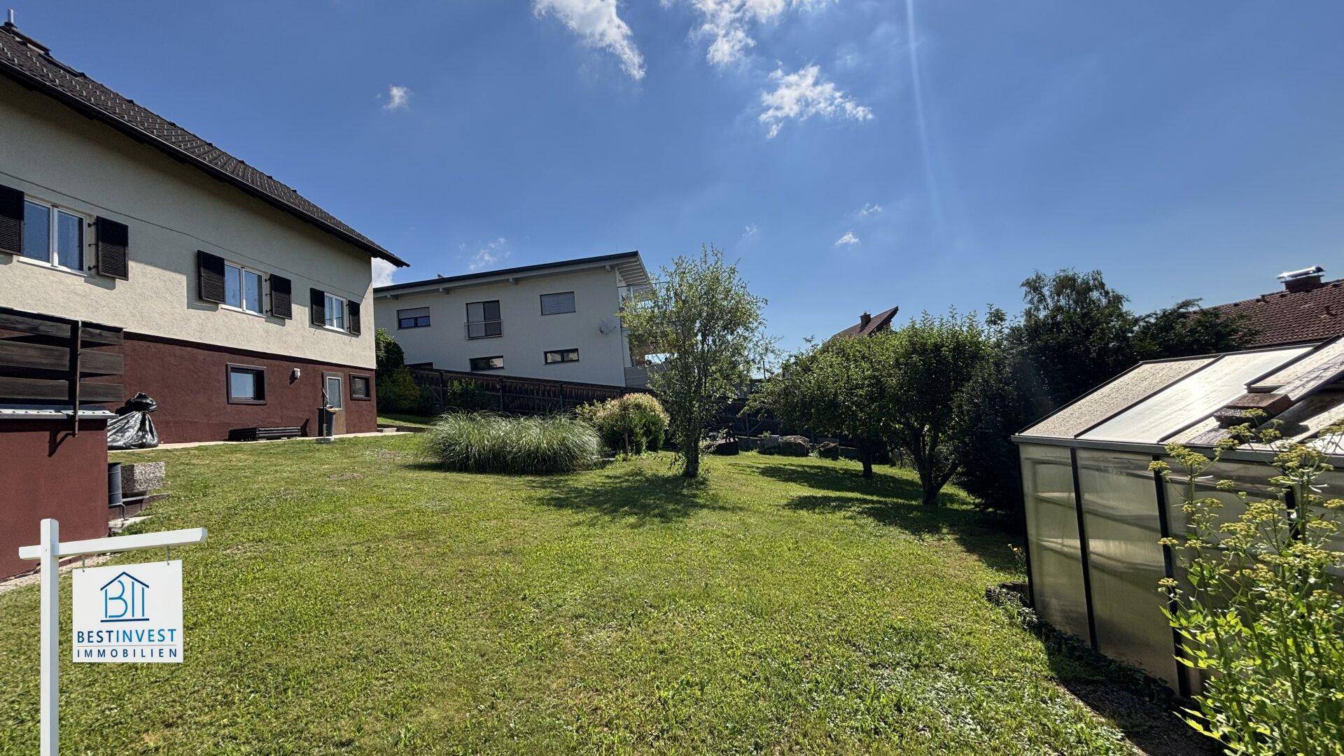 Großer Garten mit Rasenfläche, Bäumen und einem Gewächshaus im Hintergrund.