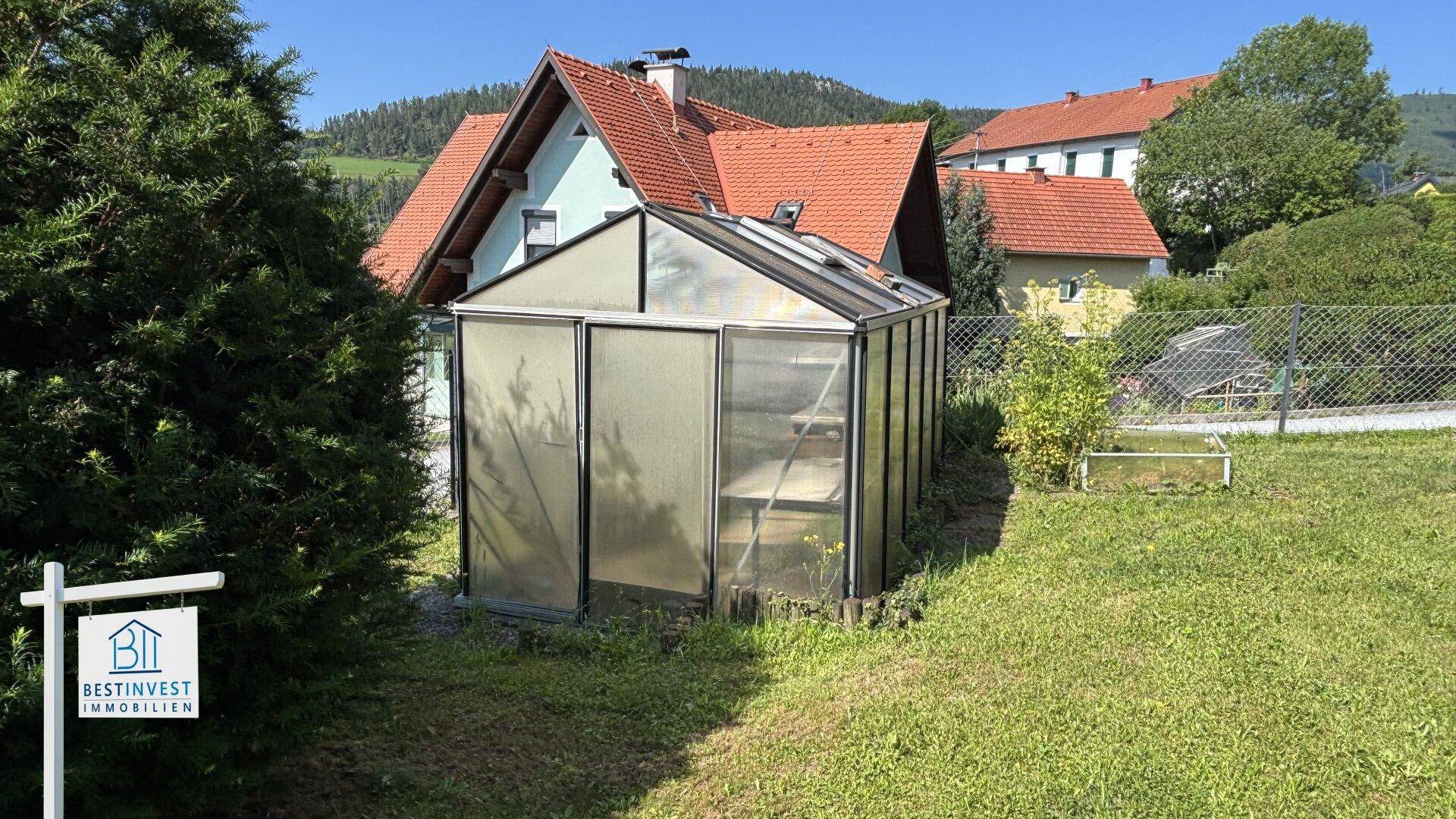 Gewächshaus im Garten mit umliegender Grünfläche und Blick auf das Wohnhaus.
