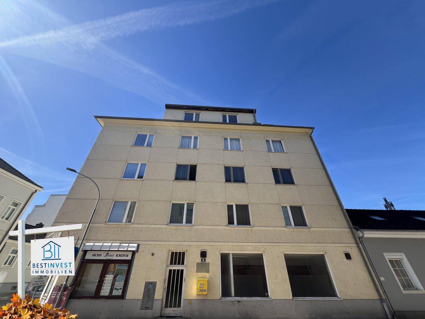 Vorderansicht eines mehrstöckigen Gebäudes mit hellgelber Fassade und Ladenlokalen im Erdgeschoss.