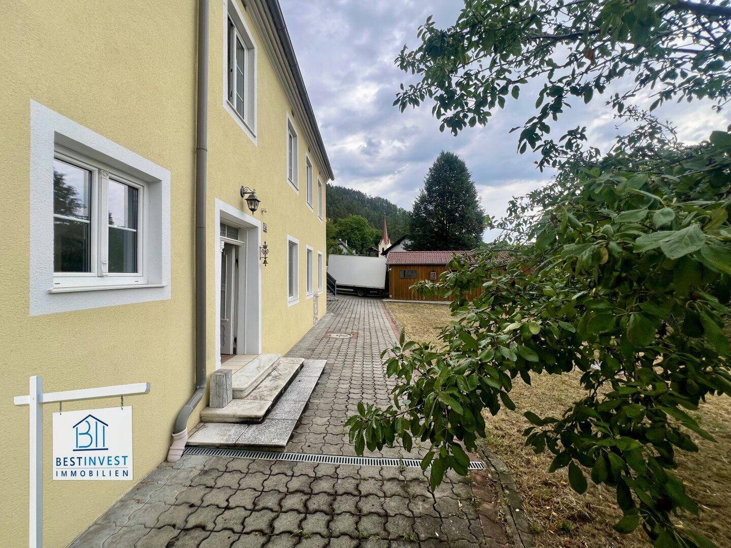Seitenansicht eines gelben Gebäudes mit gepflastertem Weg, Stufen und Bäumen im Hintergrund.