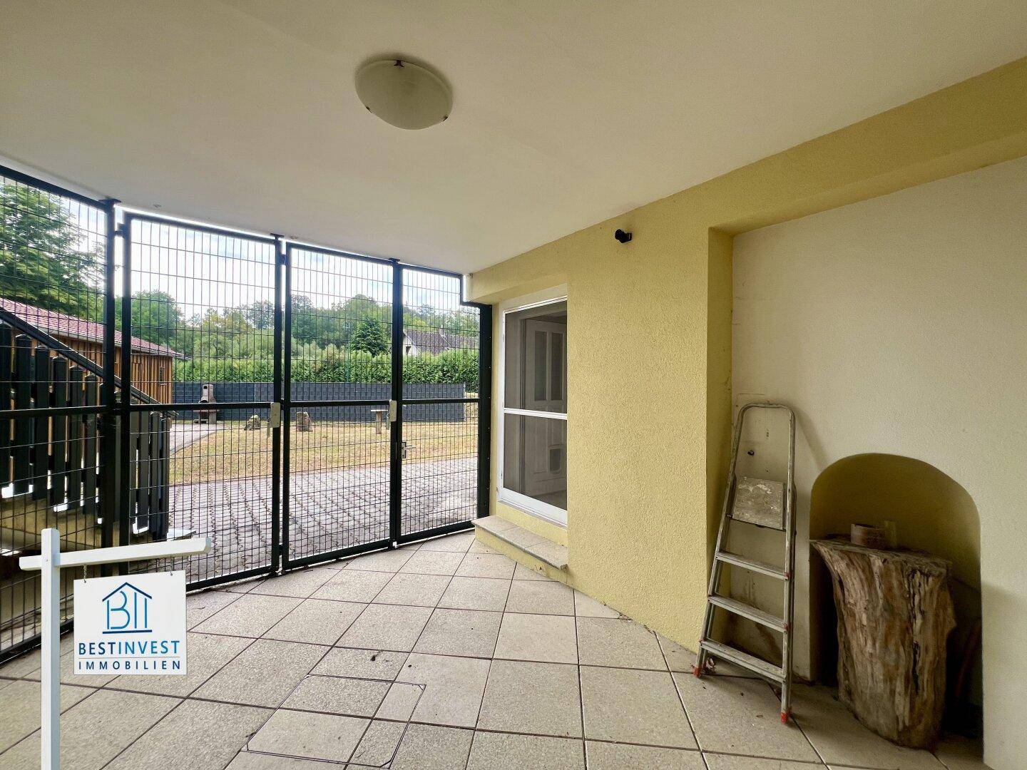 Überdachte Terrasse mit Fliesenboden, gelber Fassade und einem stabilen Metallzaun im Außenbereich.