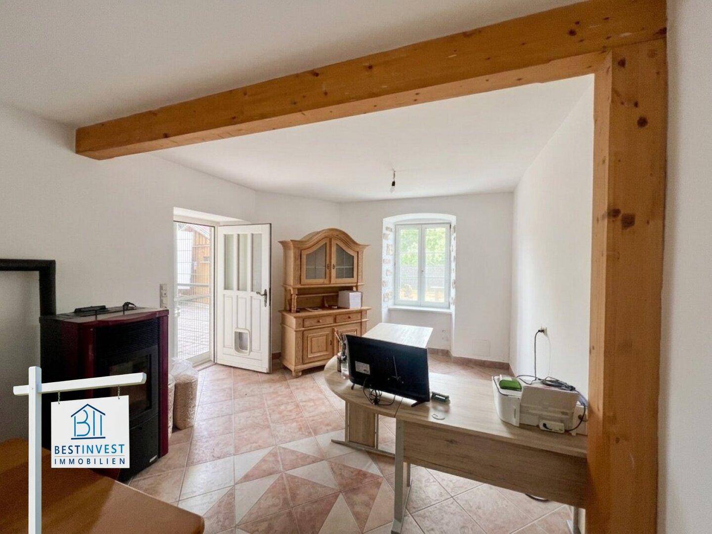 Helles Büro oder Wohnzimmer mit Schreibtisch, Holzschrank, Holzofen und sichtbarem Deckenbalken.
