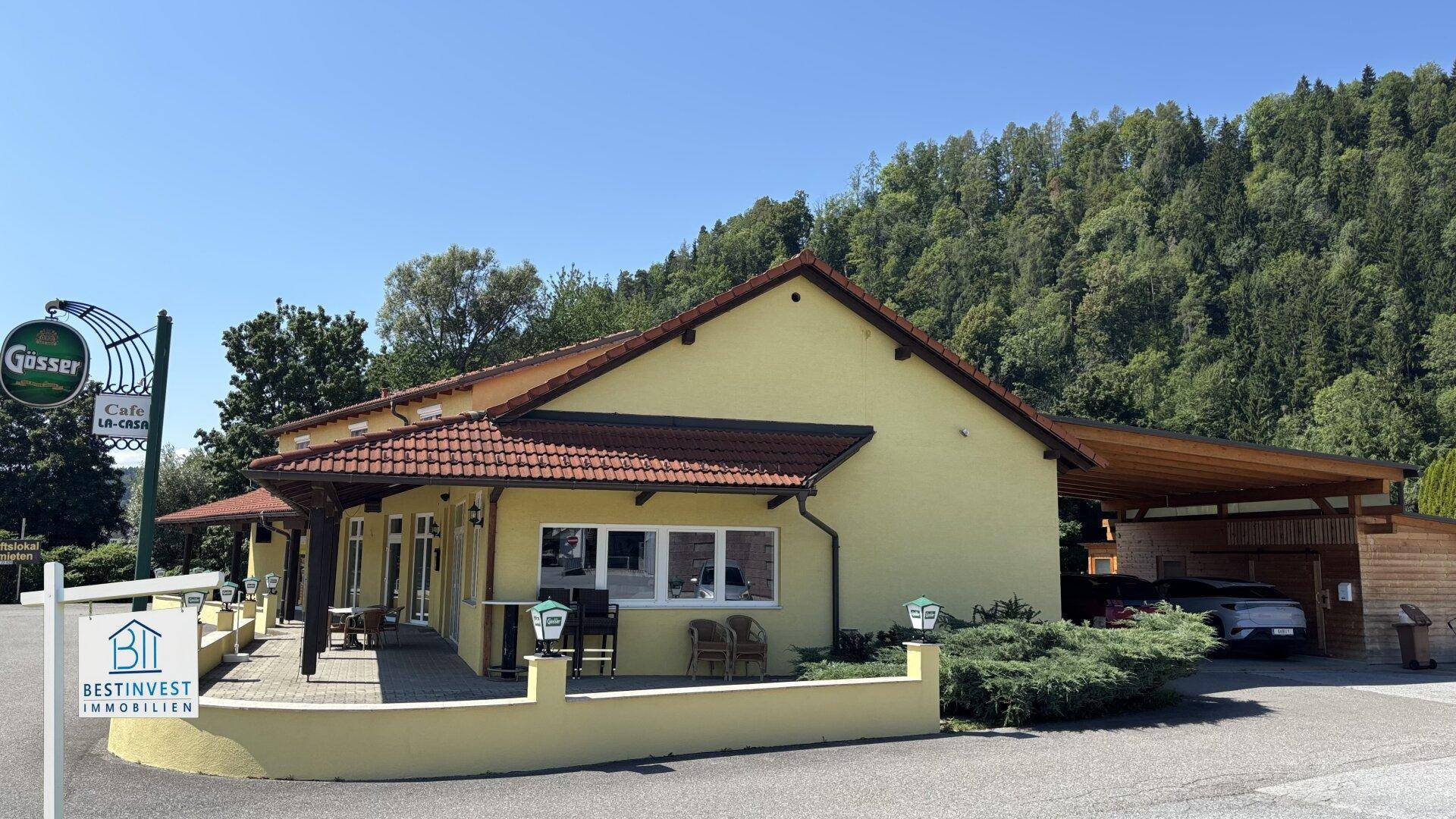 Seitenansicht des Gebäudes mit Carport und umgebender Natur unter blauem Himmel.