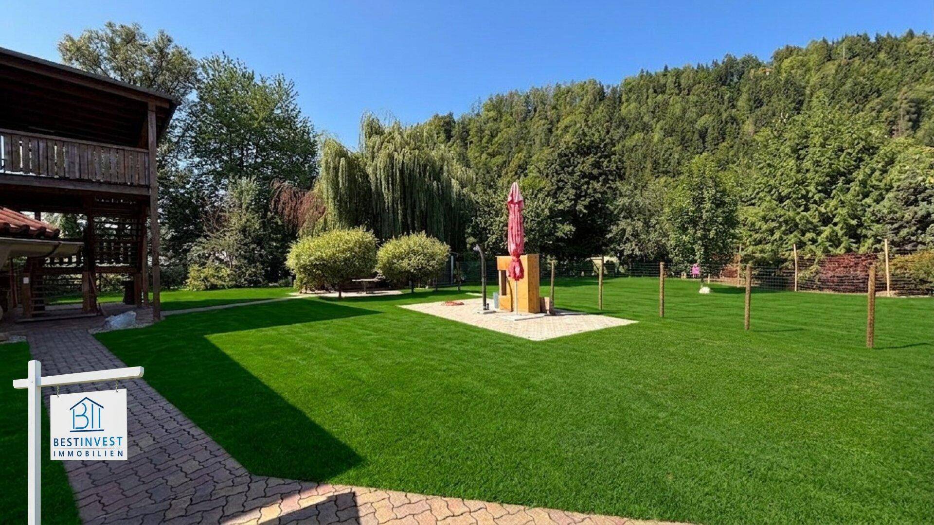 Weitläufiger Rasenbereich im Garten mit gepflasterter Fläche und Blick auf die Holzterrasse.