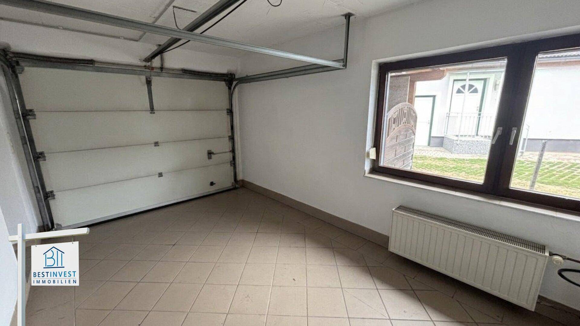 Leere Garage mit Fliesenboden, großem Fenster und Rolltor, bereit zur Nutzung.