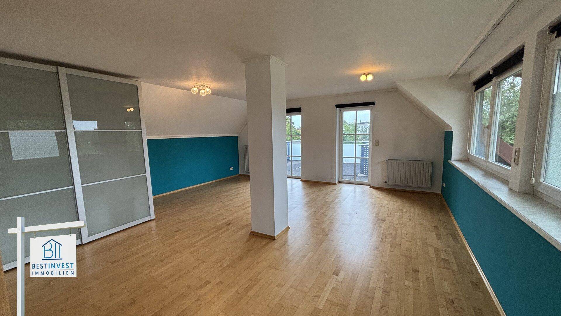 Geräumiges Dachgeschosszimmer mit Holzboden, blauer Akzentwand und Balkonzugang.