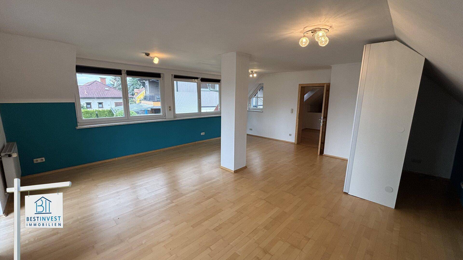 Helles Dachgeschosszimmer mit Holzboden, blauer Akzentwand und mehreren Fenstern.
