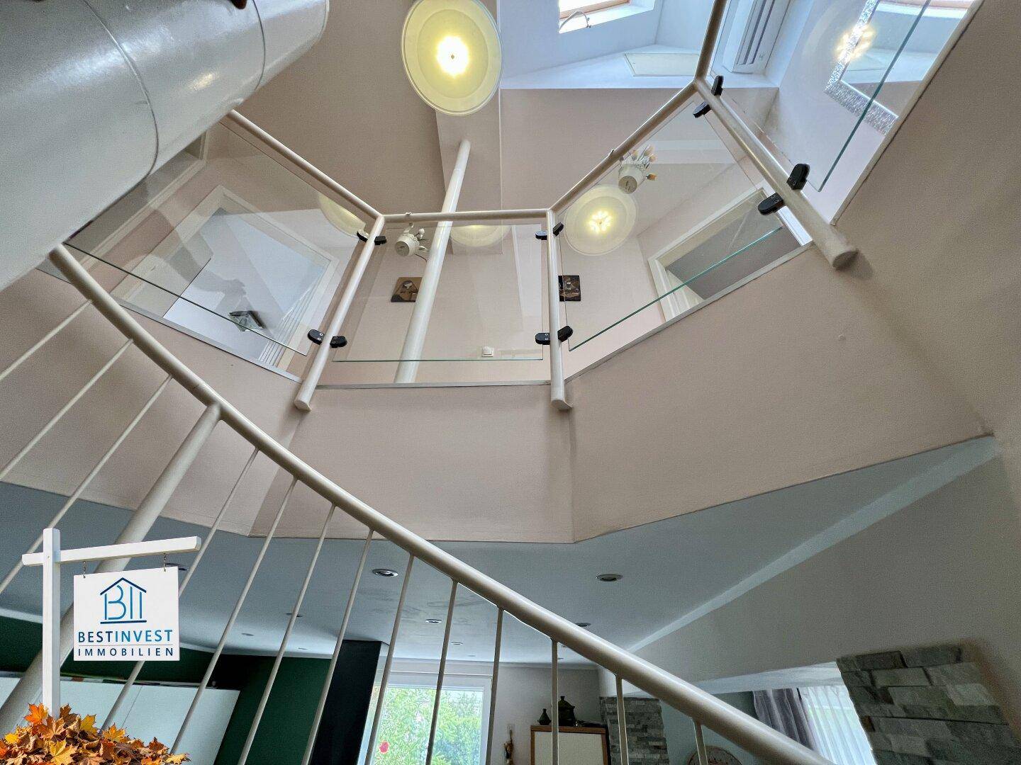 Blick nach oben auf die Wendeltreppe mit Glasgeländer und heller Deckenbeleuchtung.