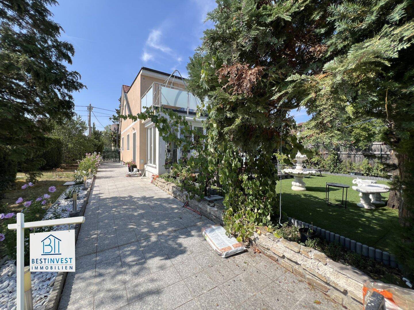 Gepflasterter Weg führt entlang des Hauses durch den grünen Garten mit Bäumen und Brunnen.
