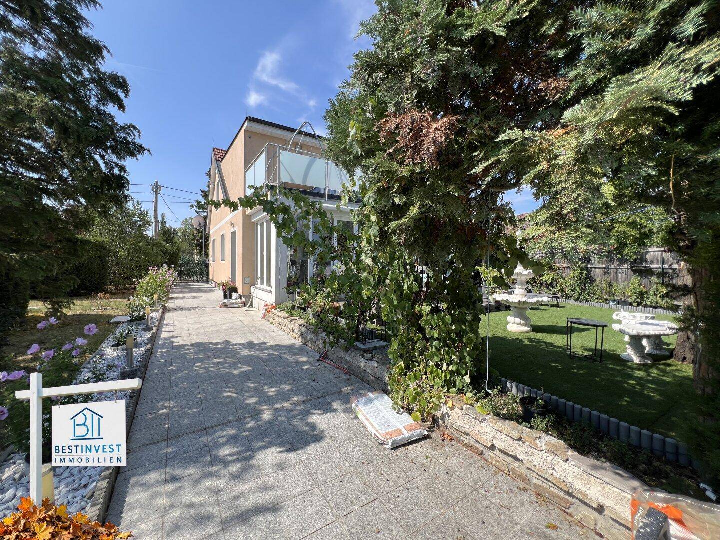 Seitenansicht des Hauses mit gepflastertem Weg, umgeben von Bäumen und einem grünen Gartenbereich.