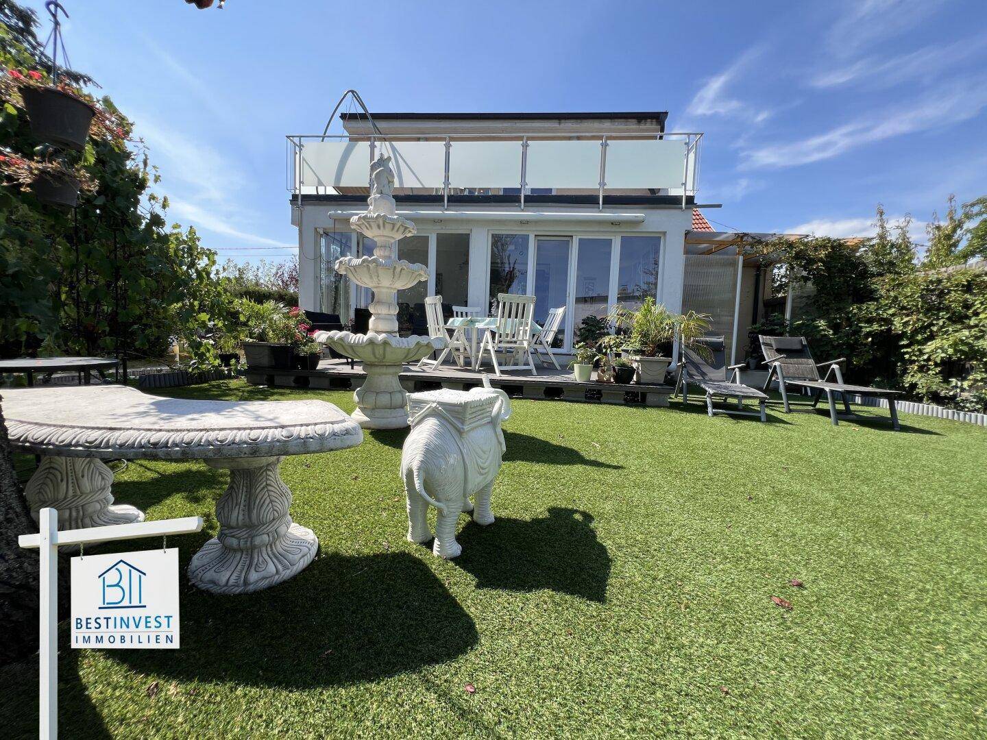 Hinterer Bereich des Hauses mit Terrasse, Kunstrasen, dekorativem Brunnen und Liegestühlen.