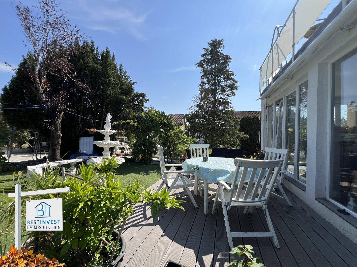 Sonnige Terrasse mit Gartenmöbeln und Blick auf den gepflegten Rasen und einen dekorativen Brunnen.
