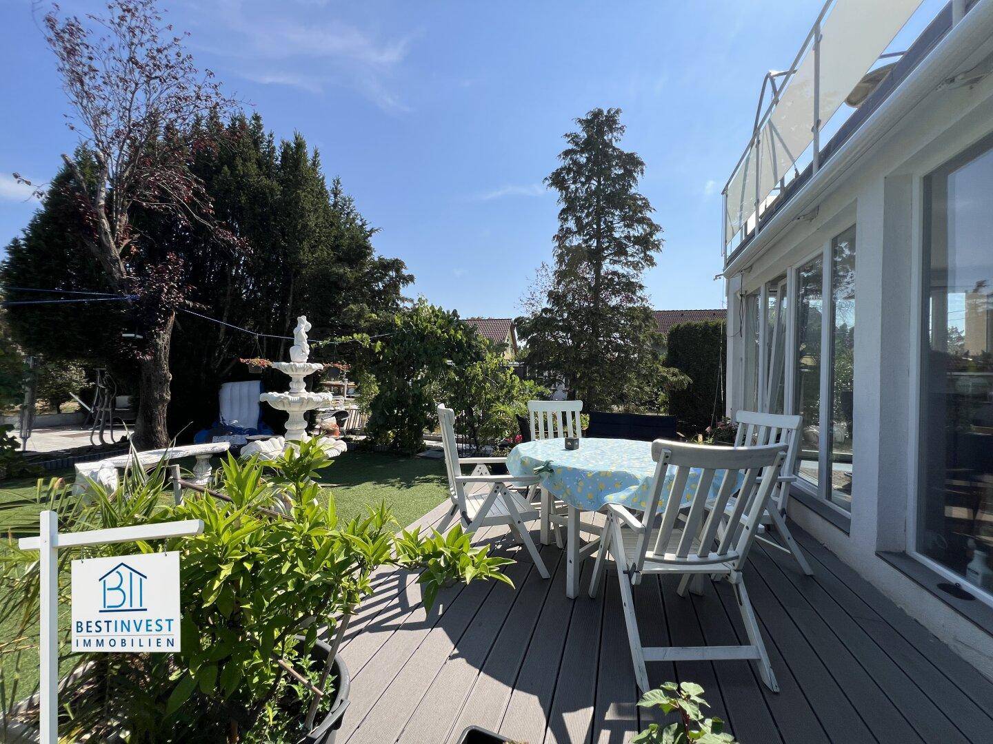 Sonnige Terrasse mit Gartenmöbeln, Rasenfläche und dekorativem Brunnen im Freien.