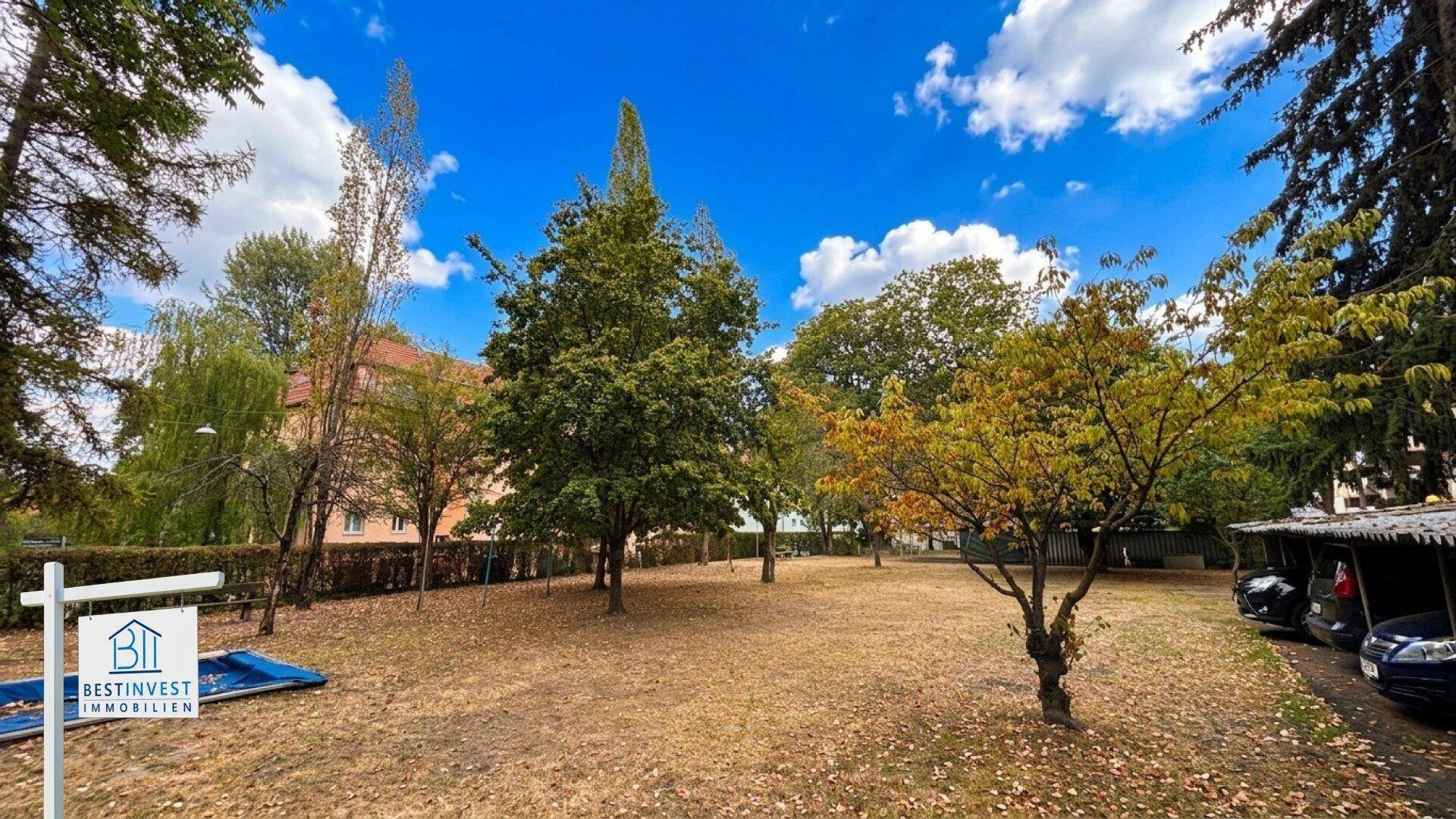 Großer Gemeinschaftsgarten mit Bäumen und überdachten Parkplätzen im Hintergrund.