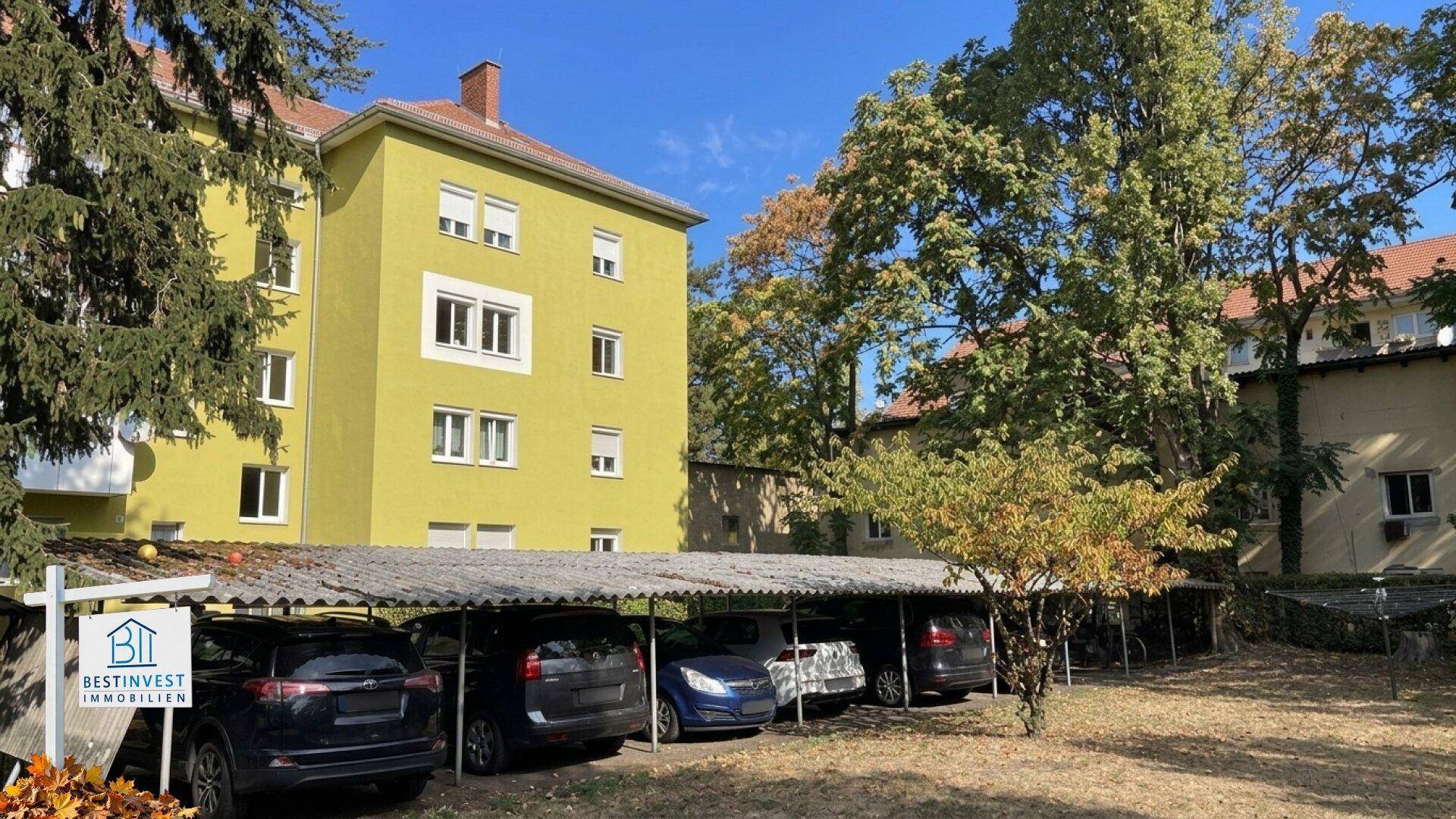 Gelbes Mehrfamilienhaus mit Carports und Bäumen im Vordergrund unter blauem Himmel.