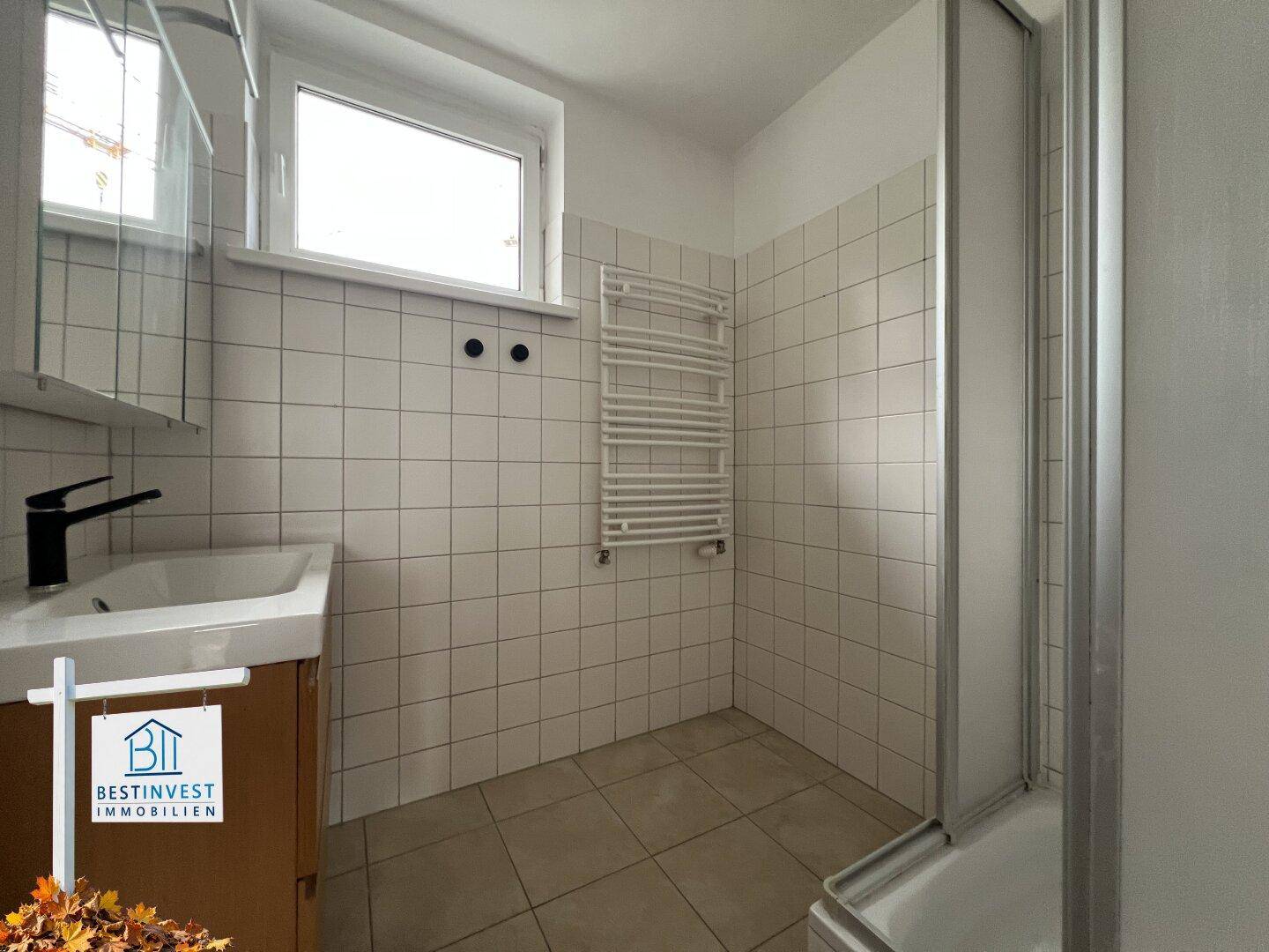 Gefliestes Badezimmer mit Dusche, Waschbecken und beheiztem Handtuchhalter, hell durch ein Fenster.