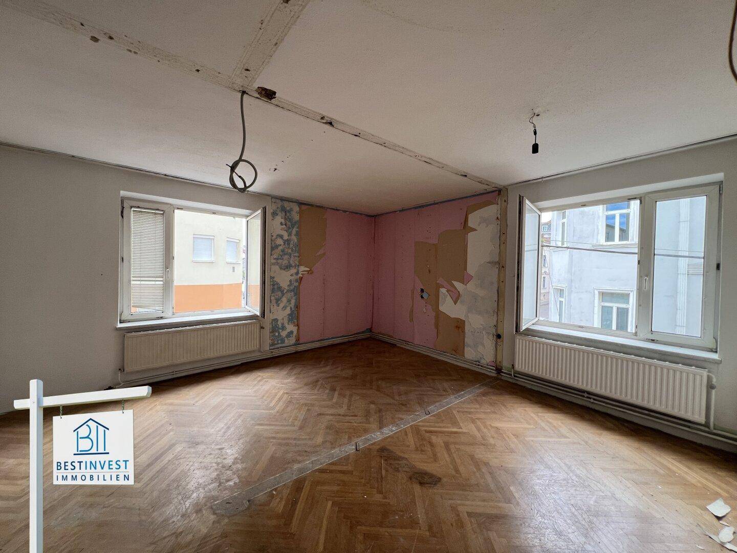 Renovierungsbedürftiges Zimmer mit alten Tapetenresten, Parkettboden und zwei Fenstern.