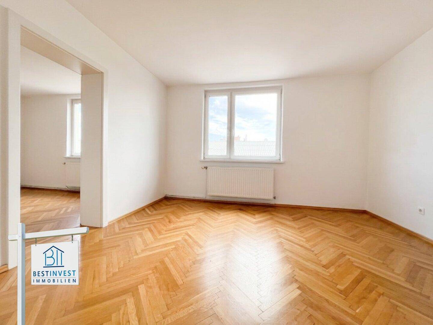Geräumiges Zimmer mit Fischgrätparkettboden und großen Fenstern, die viel Tageslicht hereinlassen.