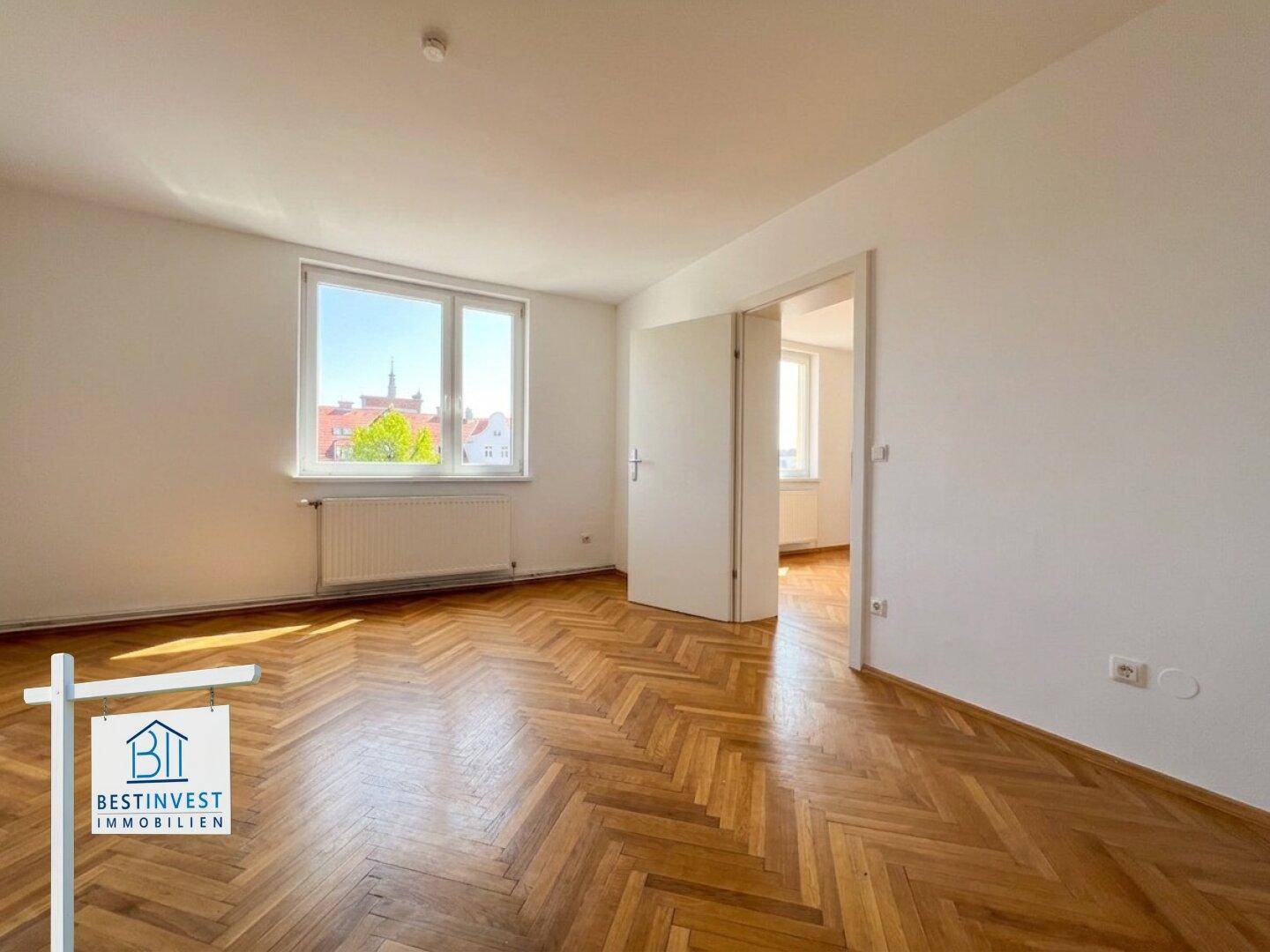 Helles Zimmer mit klassischem Fischgrätparkett und Blick aus dem Fenster auf die grüne Umgebung.