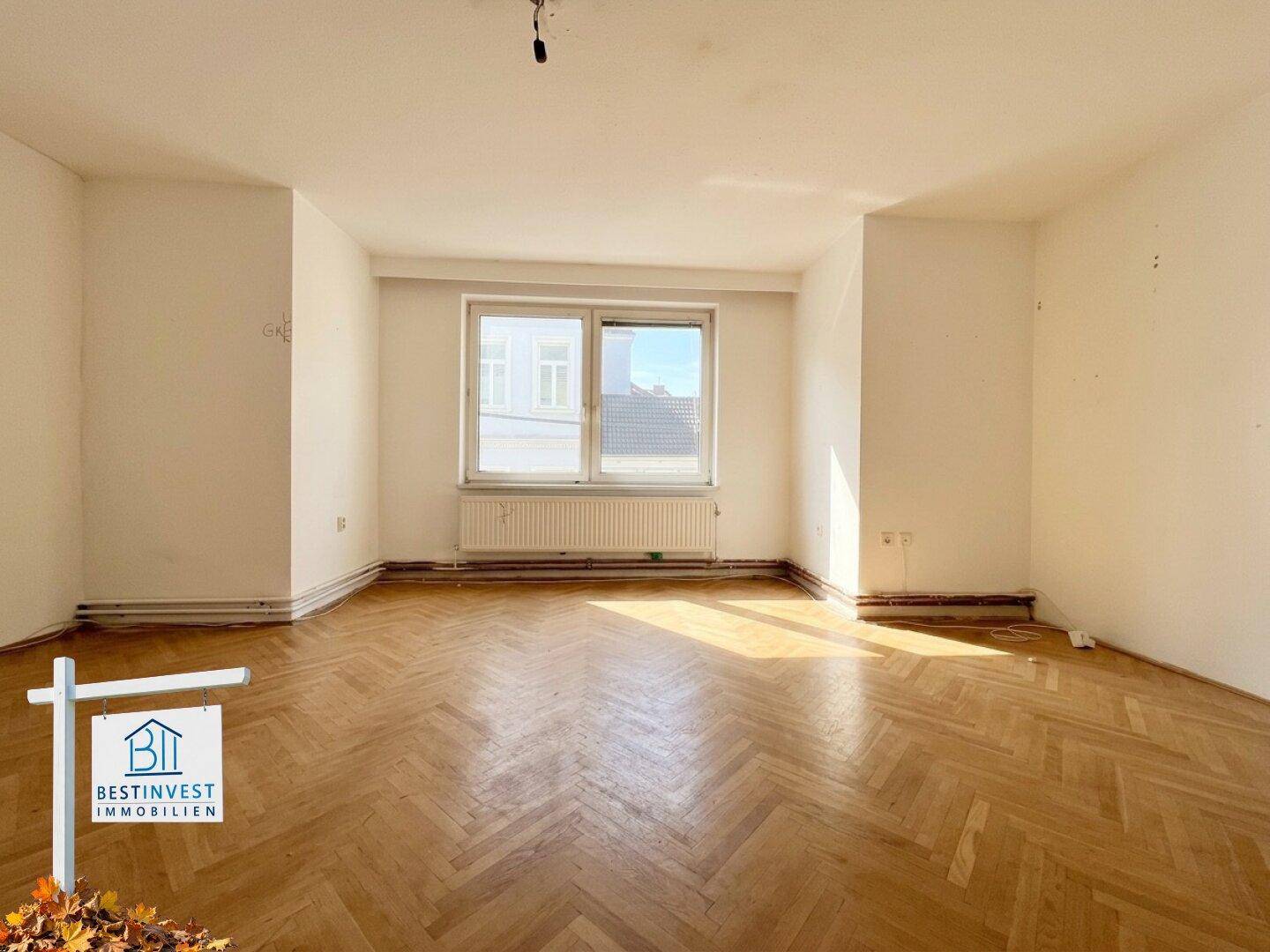 Helles, leeres Zimmer mit Fischgrätparkett und großem Fenster, das viel Sonnenlicht einfängt.