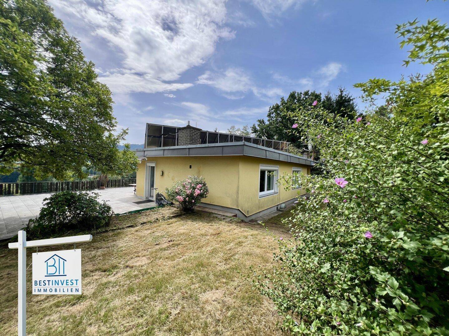 Seitenansicht des gelben Hauses mit Gartenbereich und Bäumen unter blauem Himmel.