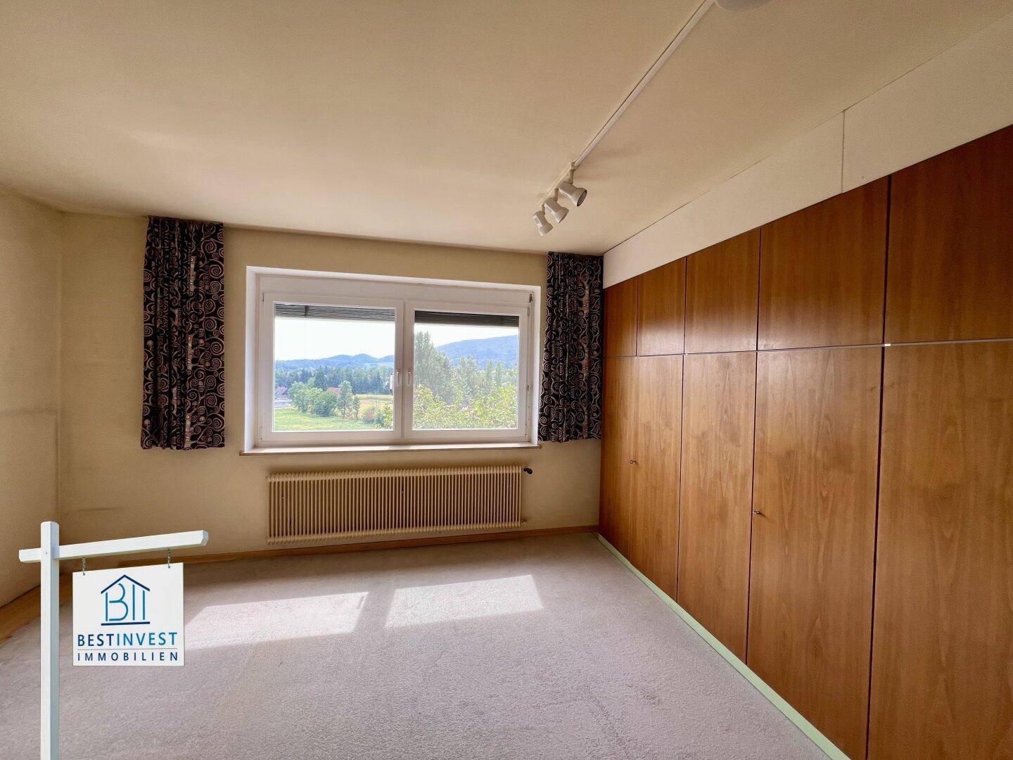 Helles Schlafzimmer mit großem Fenster, Einbauschränken aus Holz und Blick ins Grüne.