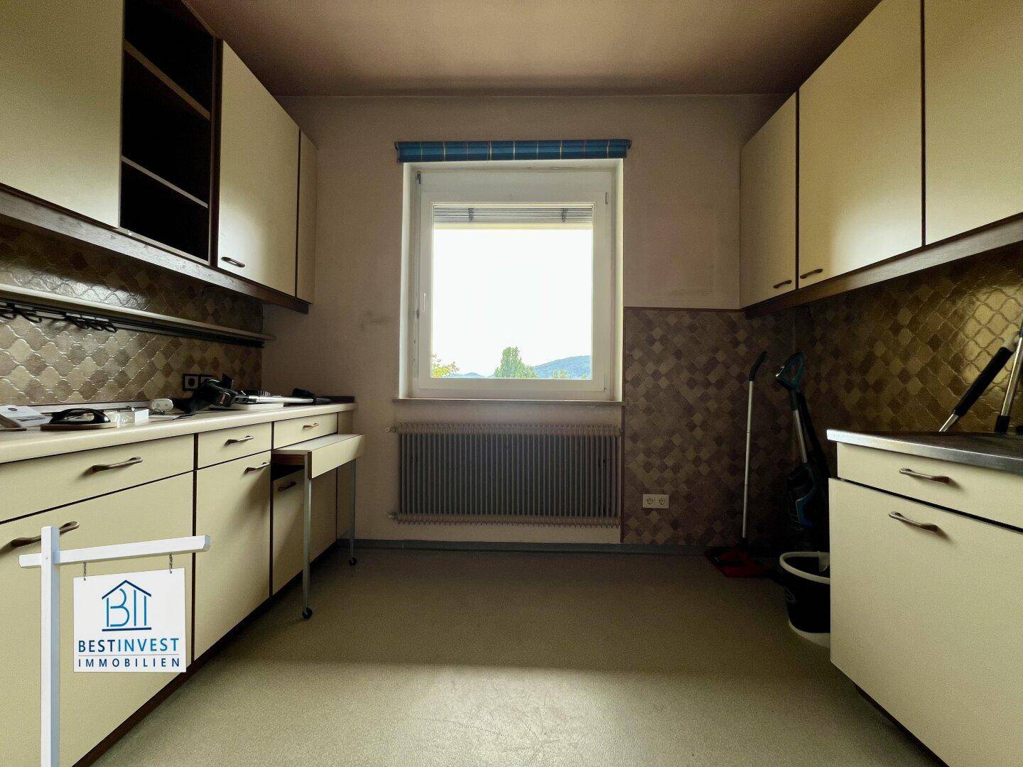 Ältere Küche mit beigen Schränken, Fenster und Heizkörper, die eine Renovierung benötigt.