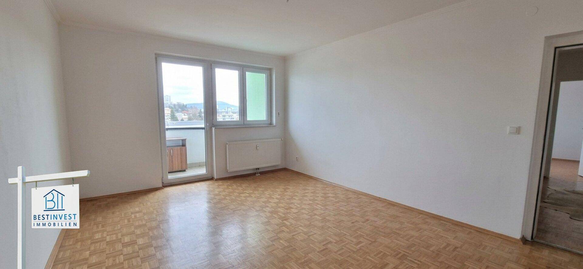 Geräumiges, leeres Wohnzimmer mit Parkettboden und Zugang zu einem kleinen Balkon.