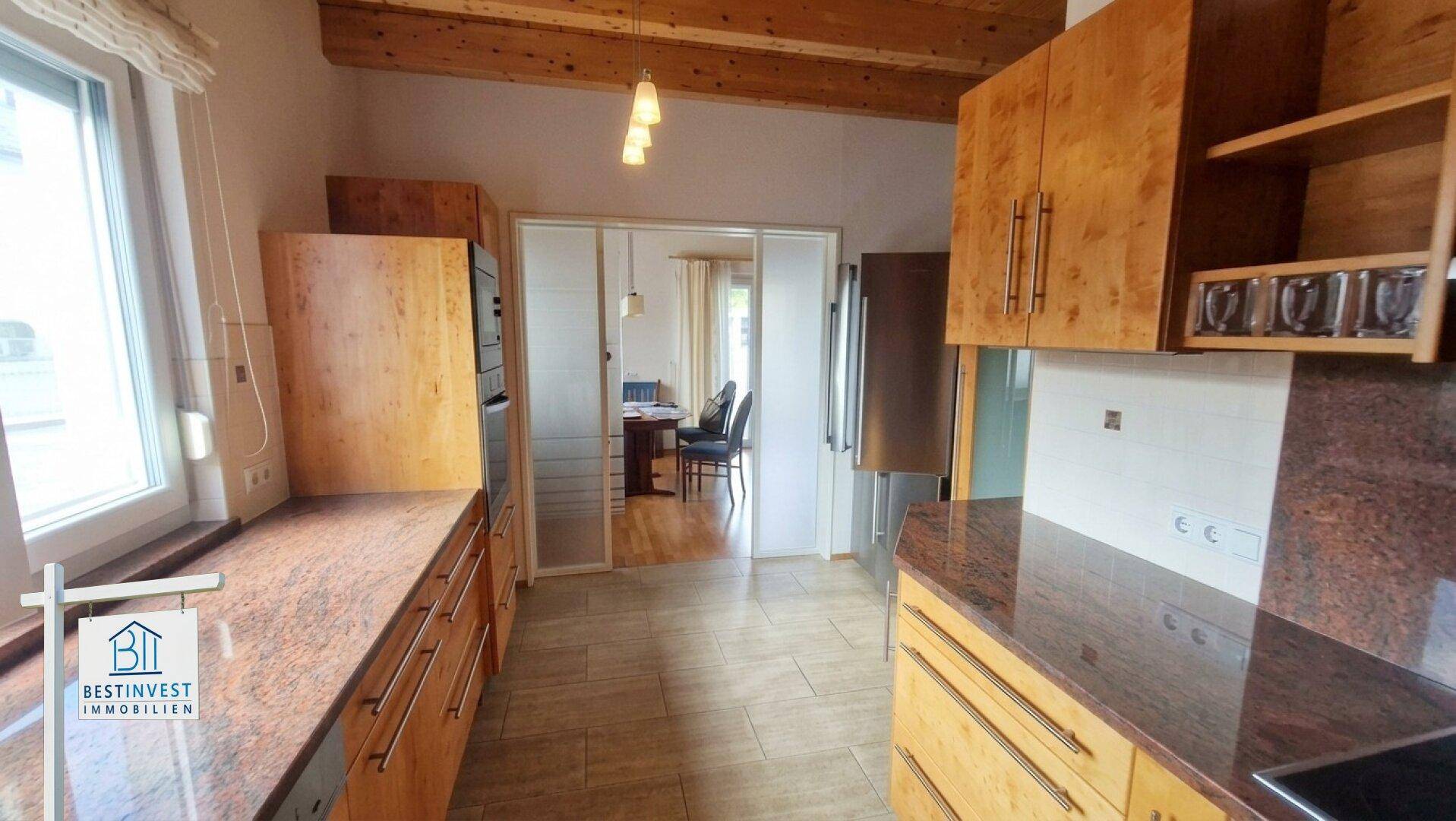 Küche mit Holzschränken, Granitarbeitsplatten und Fliesenboden, mit Blick in den Essbereich.