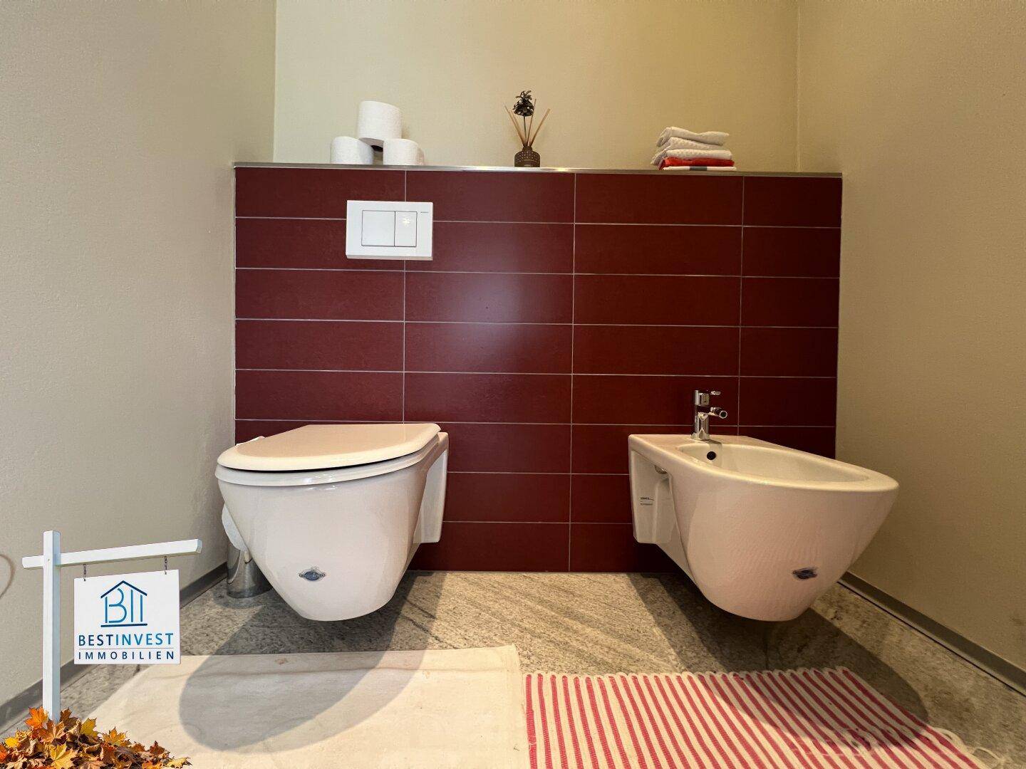 Modernes WC mit wandhängender Toilette und Bidet, akzentuiert durch rote Fliesen.