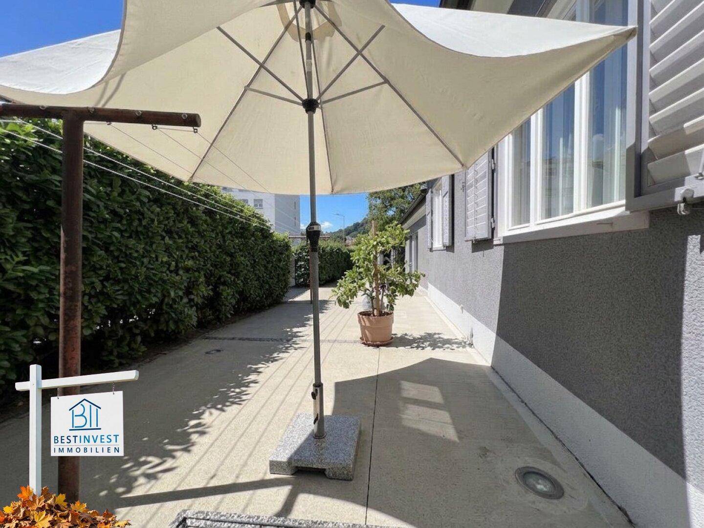 Langer, gepflasterter Weg entlang des Hauses mit hoher Hecke und großem Sonnenschirm.