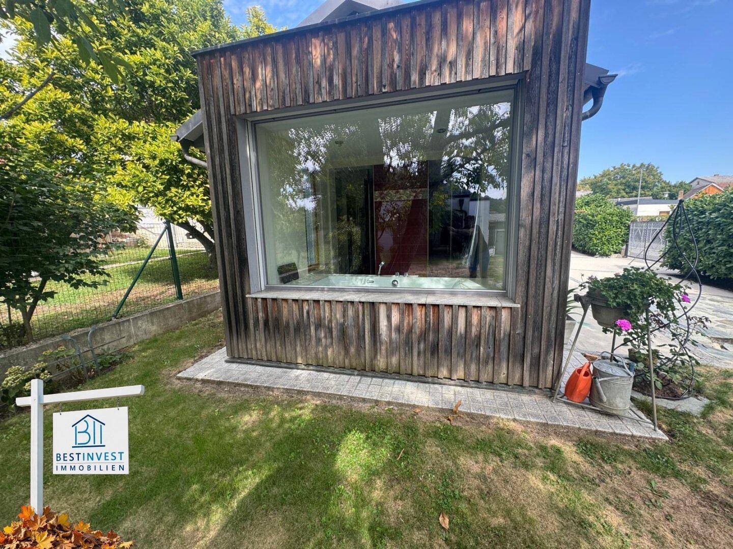 Holzverkleidetes Gebäude mit großem Fenster und direktem Zugang zum grünen Gartenbereich.