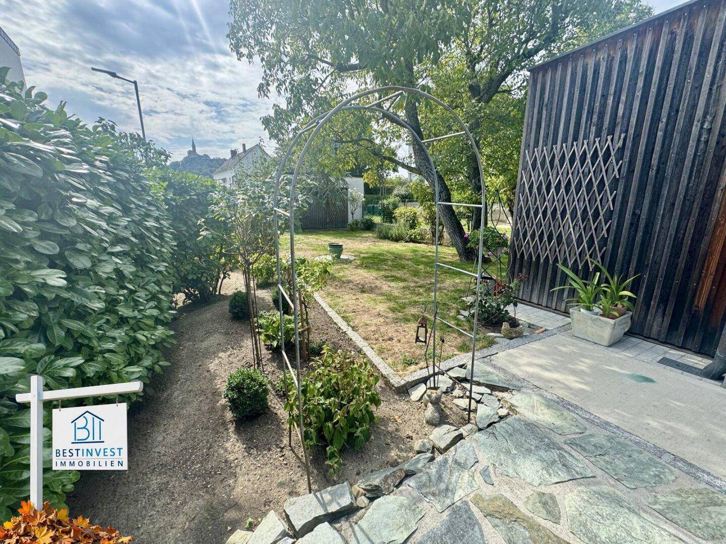 Idyllischer Gartenweg mit Rankhilfe und grüner Bepflanzung neben einer Holzfassade.