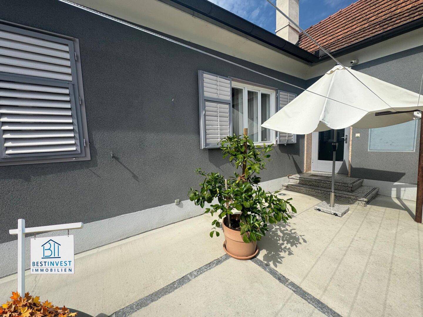 Gemütlicher Terrassenbereich vor der Hausfassade mit Sonnenschirm und Topfpflanze.