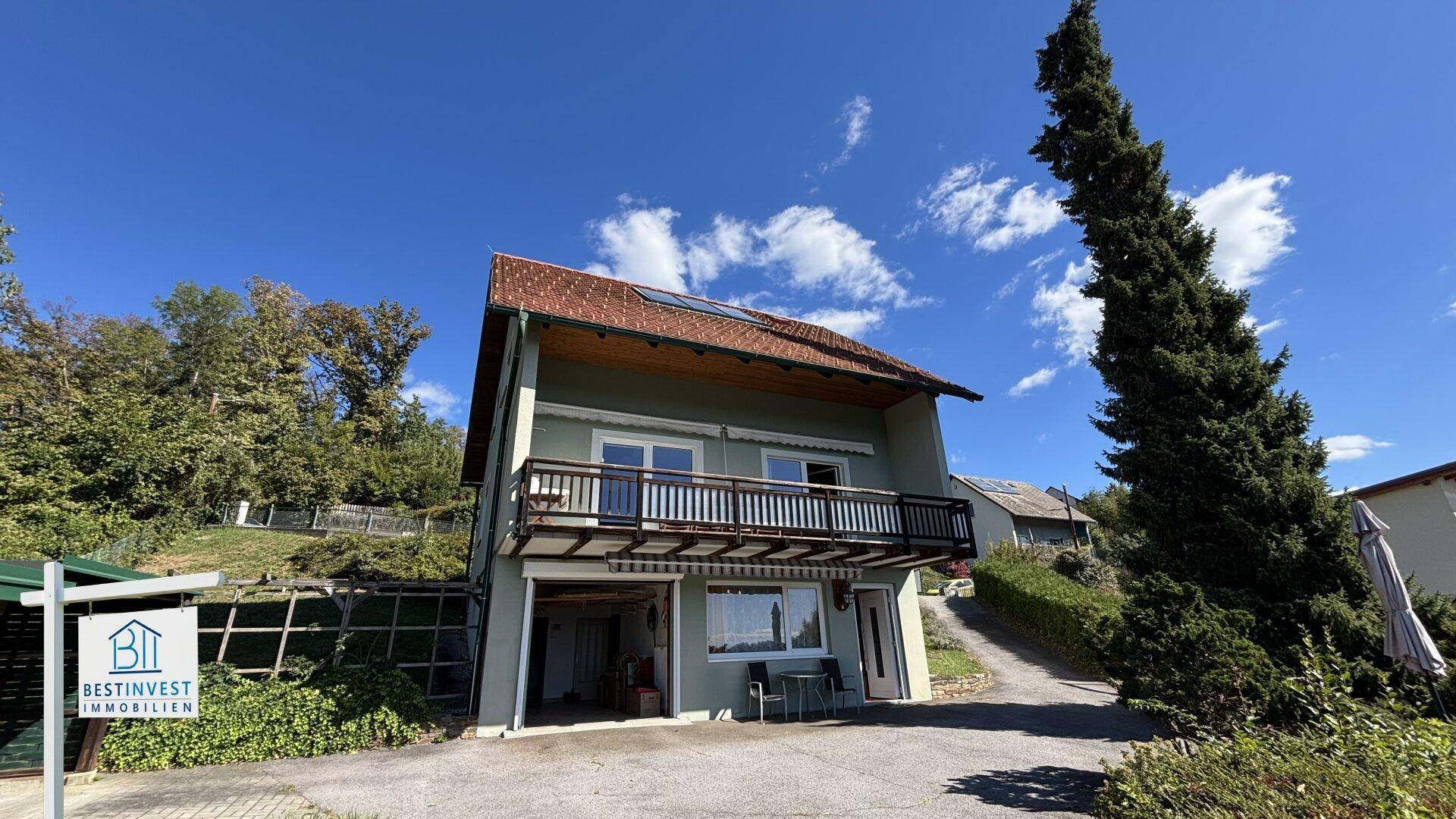 Wohnhaus mit Balkon und Zufahrt, eingebettet in eine grüne Umgebung unter blauem Himmel.