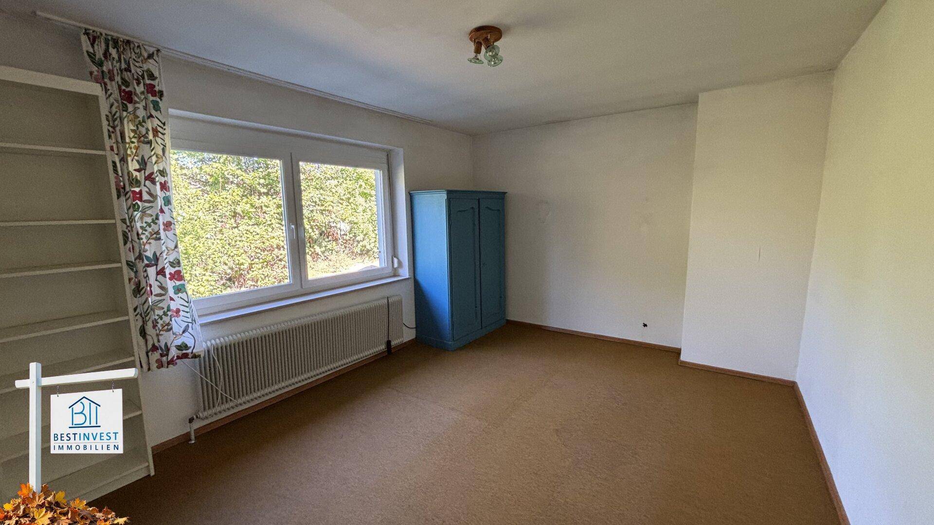 Leeres Zimmer mit Teppichboden, Fenster und einem blauen Kleiderschrank.