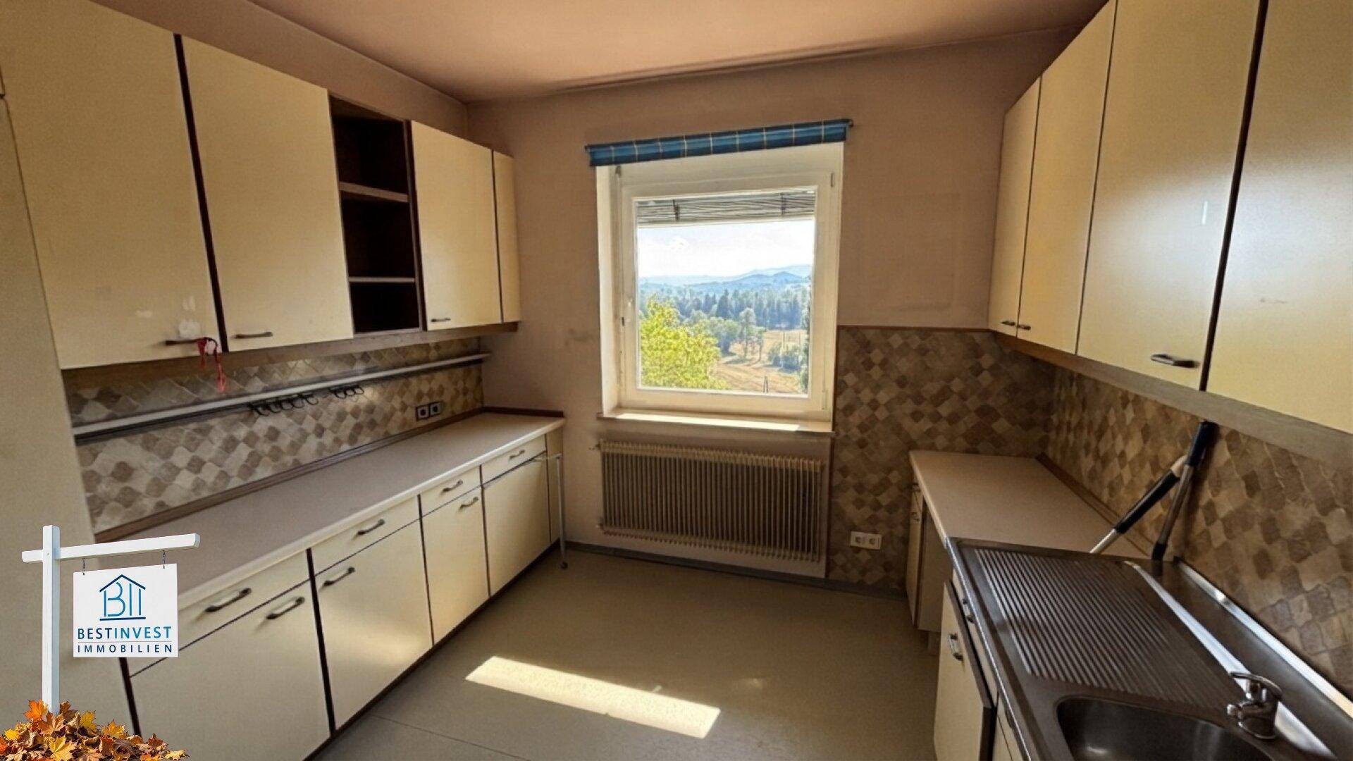 Ältere Küche mit beigen Schränken, gemustertem Fliesenspiegel und Fensterblick.