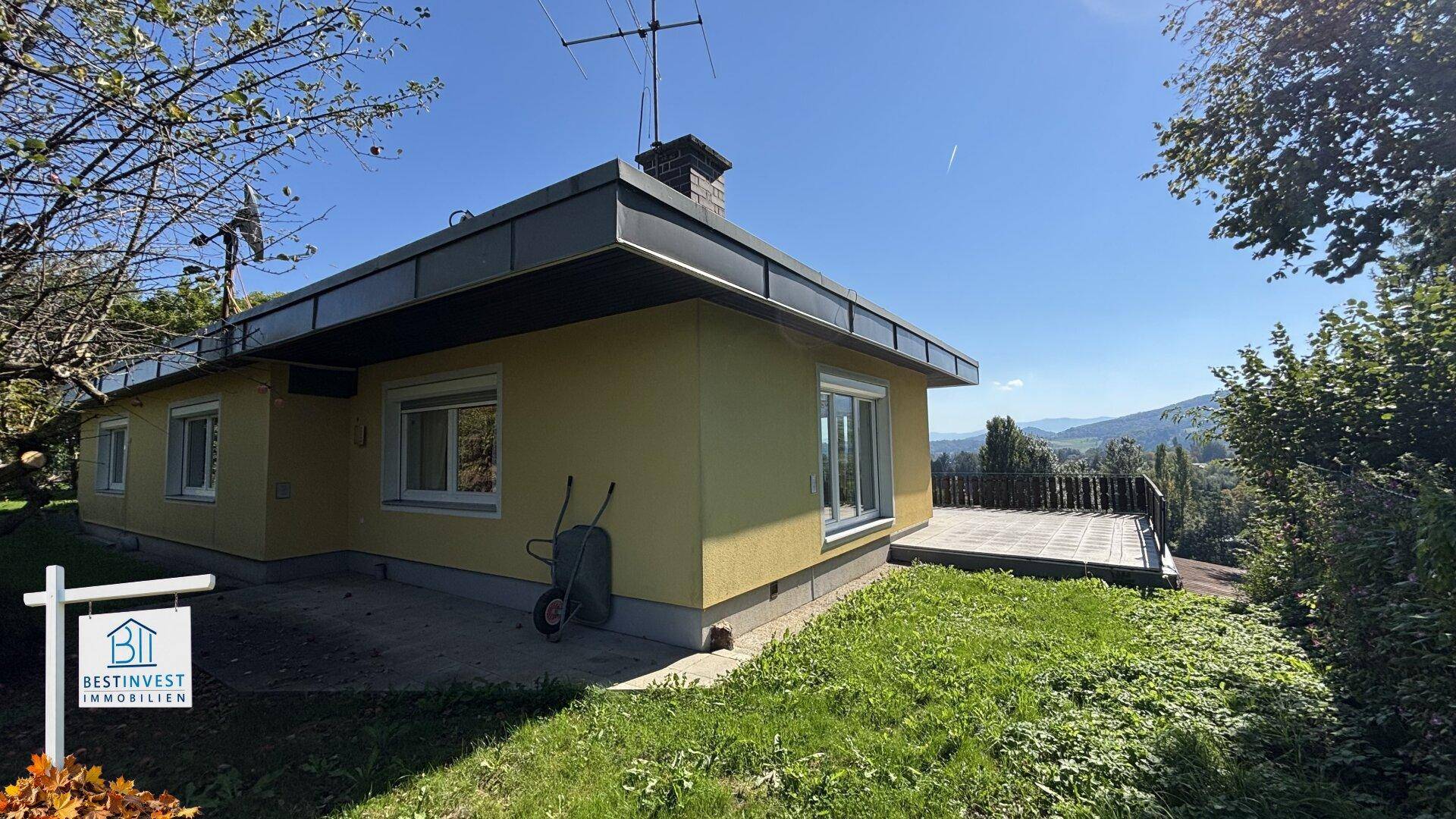 Seitliche Ansicht des gelben Hauses mit einer gepflasterten Terrasse und Rasenfläche unter blauem Himmel.