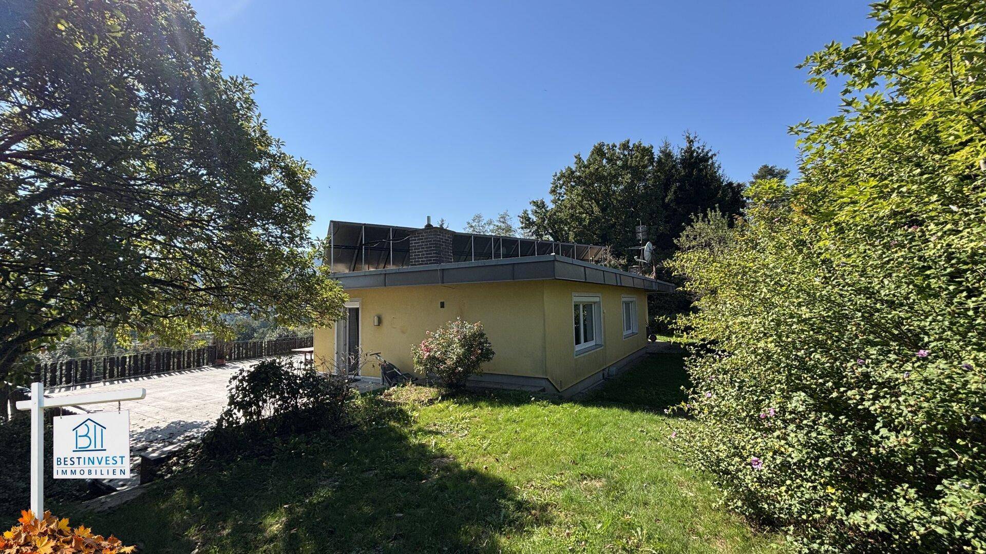 Rückansicht des gelben Hauses mit großer Terrasse, Gartenbereich und üppiger Vegetation unter blauem Himmel.