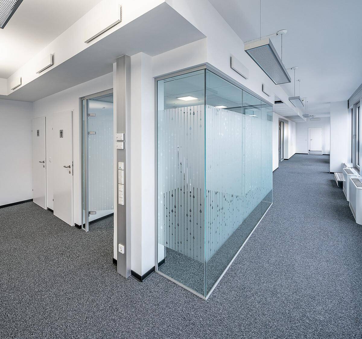 Moderner Büroflur mit grauem Teppichboden, weißen Wänden und Glasabtrennungen mit Muster.