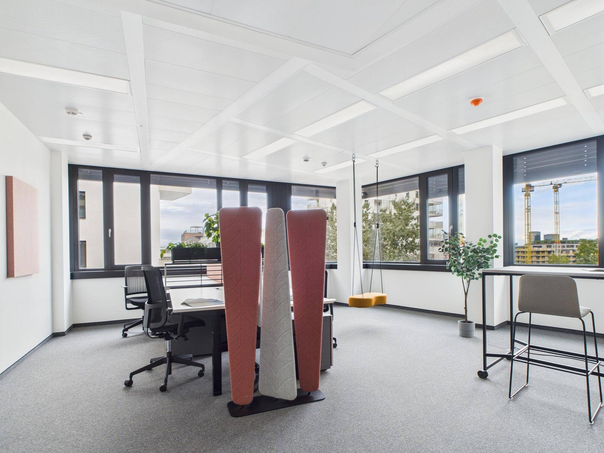 Großes, helles Büro mit mehreren Arbeitsplätzen, Teppichboden und großen Fenstern mit Stadtblick und Kränen.