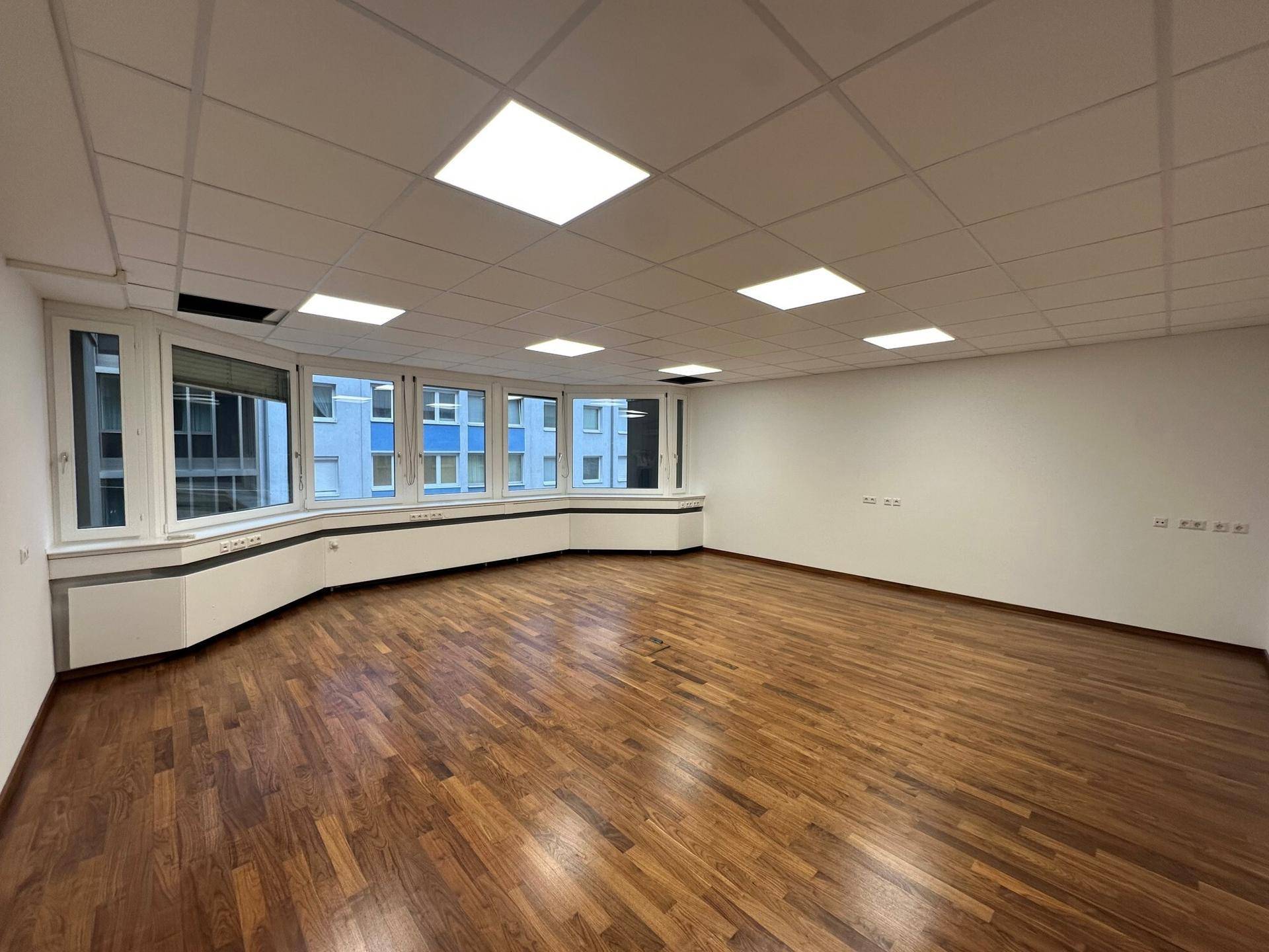 Heller Büroraum mit dunklem Holzboden und großen Fenstern mit Blick auf umliegende Gebäude.