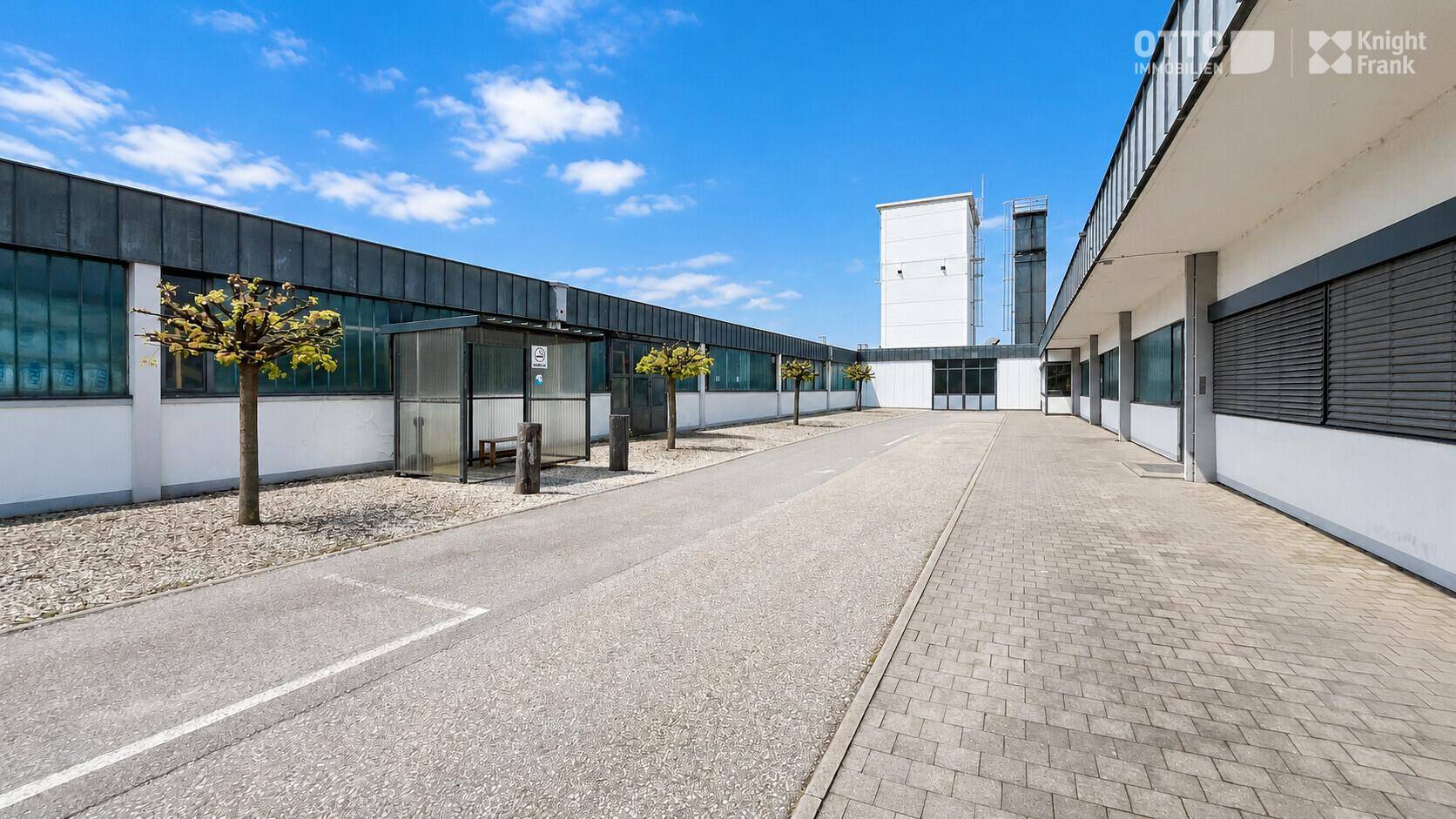 Gebäudefassade eines Gewerbekomplexes mit gepflastertem Weg, kleinen Bäumen und einer Bushaltestelle.