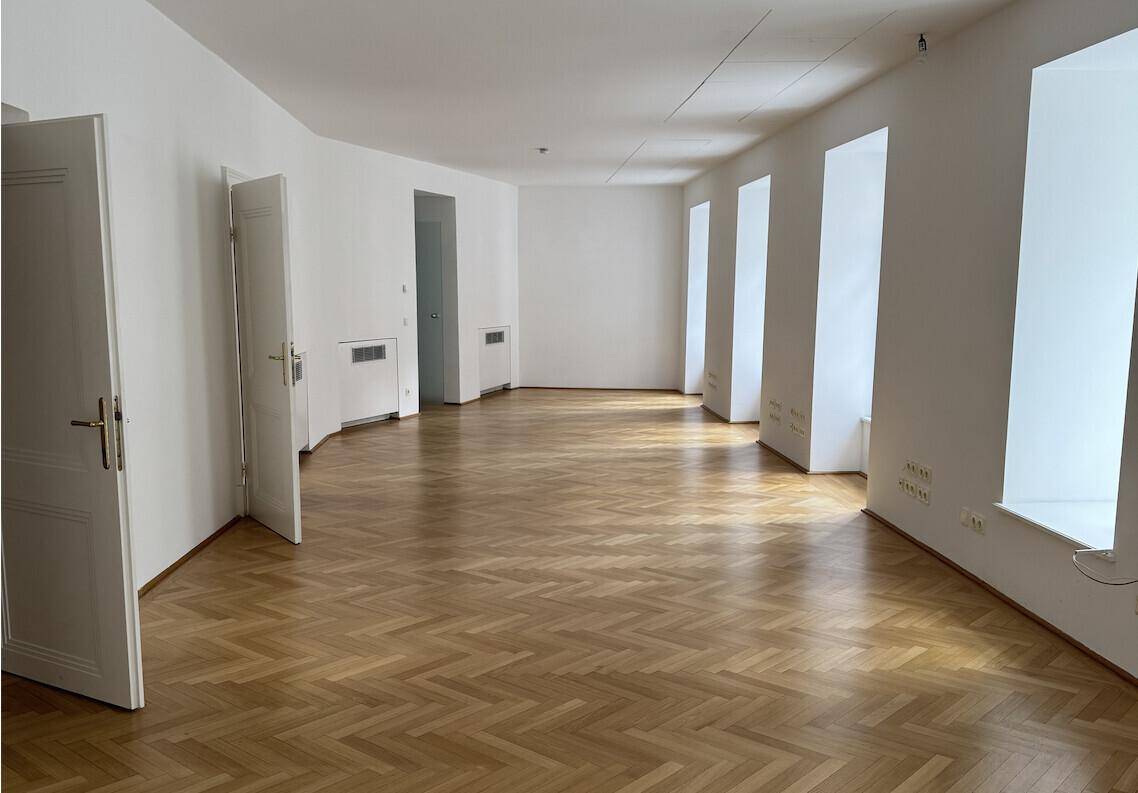 Geräumiger, leerer Raum mit mehreren Fenstern und elegantem Fischgrätparkett, ideal für ein Großraumbüro.