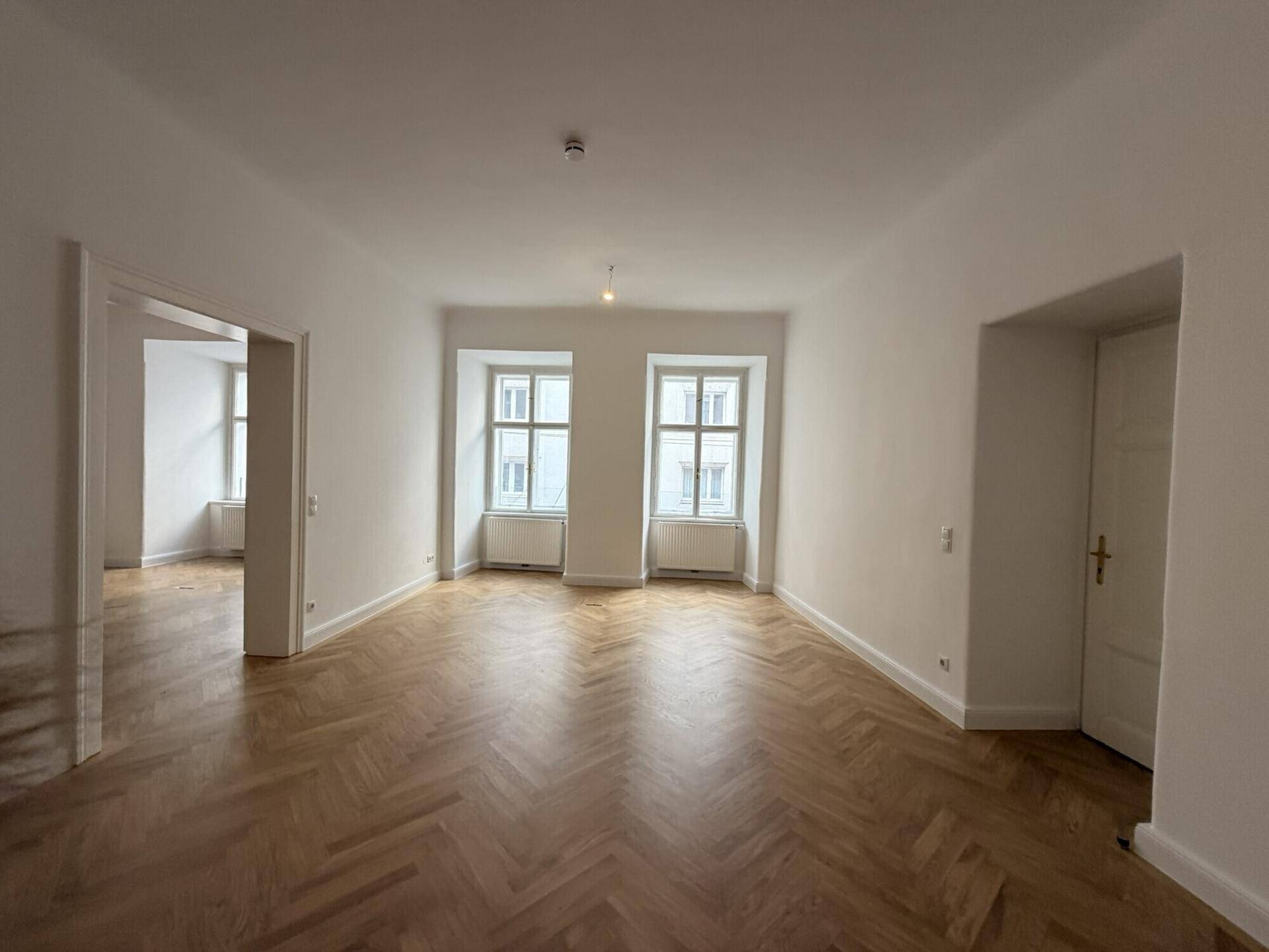 Heller Wohnraum mit Fischgrätparkett und zwei Fenstern, die den Blick nach draußen freigeben.