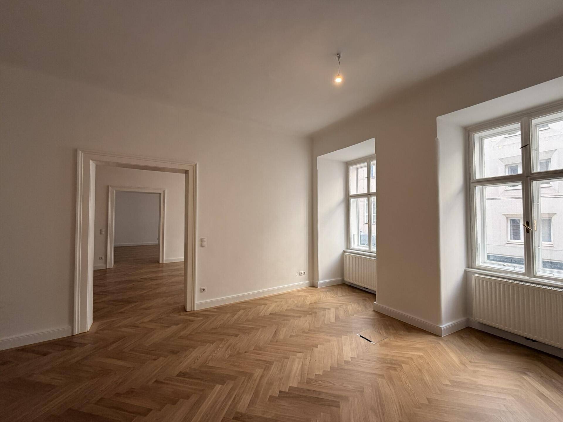 Geräumiger Wohnbereich mit Fischgrätparkett und großen Fenstern, die eine helle und einladende Atmosphäre schaffen.