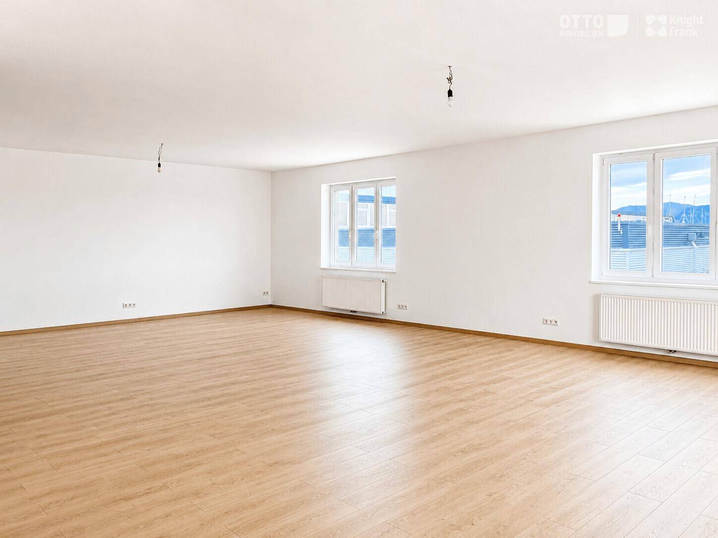Geräumiger Büroraum mit Laminatboden, weißen Wänden und großen Fenstern mit Heizkörpern.