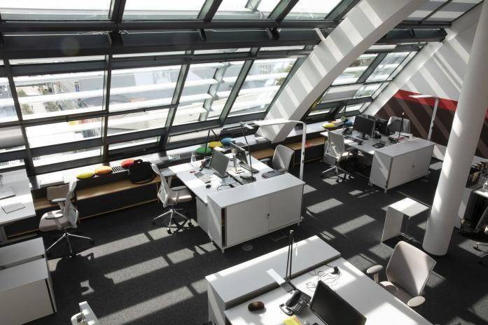 Offener Bürobereich mit modernen Arbeitsplätzen, Teppichboden und schrägen Glasfenstern, die viel Licht spenden.