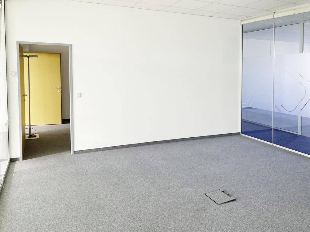 Geräumiger Büroraum mit grauem Teppichboden, weißen Wänden und einer gelben Tür.
