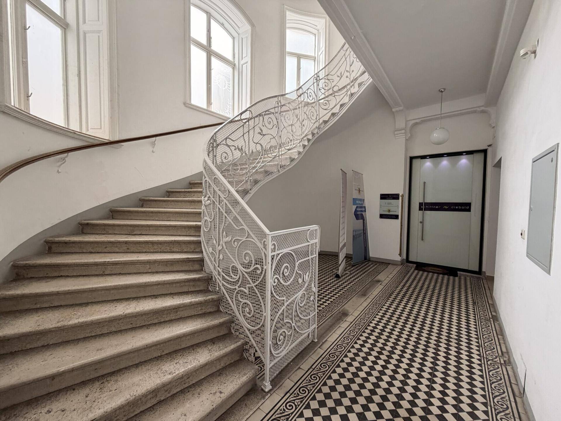 Elegantes Treppenhaus mit geschwungener Steintreppe, kunstvollem Geländer und Schachbrettmuster-Fliesenboden.